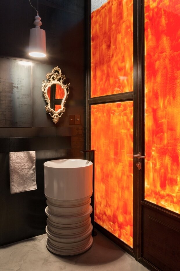 В санузле использованы различные материалы: глянцевая мелкоформатная плитка, черный металл и частично закрашенная оранжевая стеклянная перегородка, позволяющая проникать солнечному свету в ванную комнату.