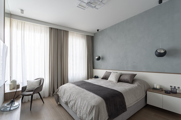 Спальня выполнена в белом цвете с акцентной стеной из микроцемента серо-голубого цвета и деревянными элементами.